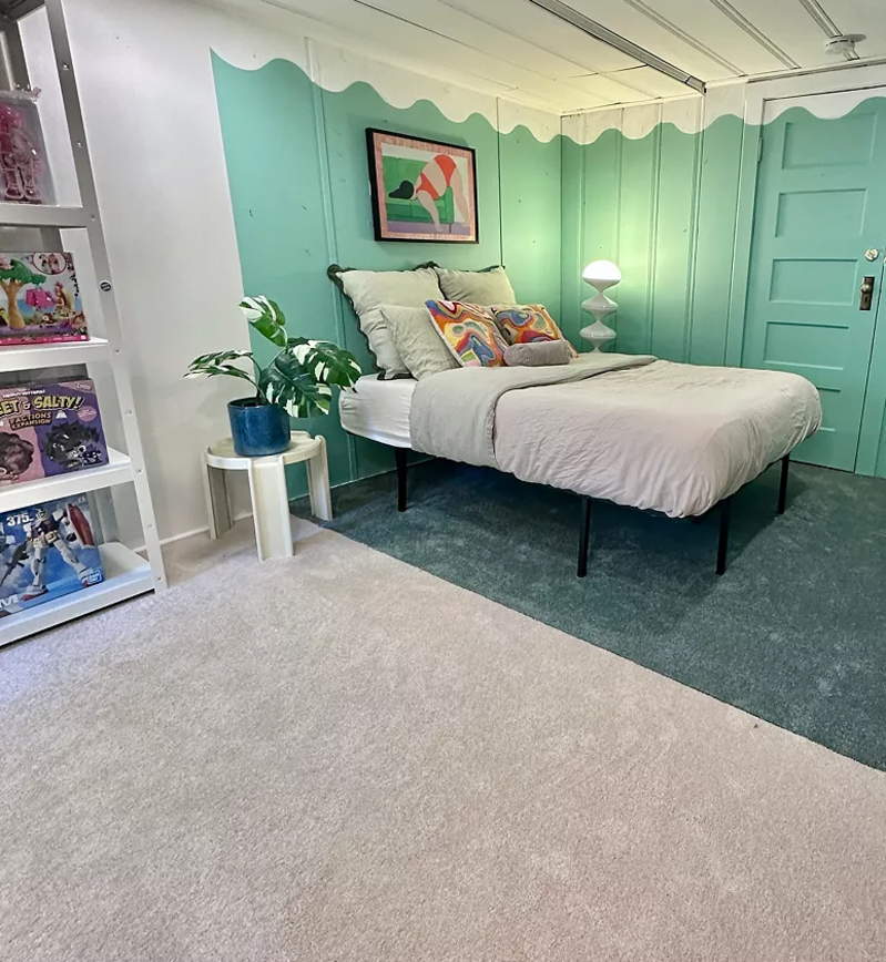 A basement room split into a sleeping area and a work area by two colors of carpet.